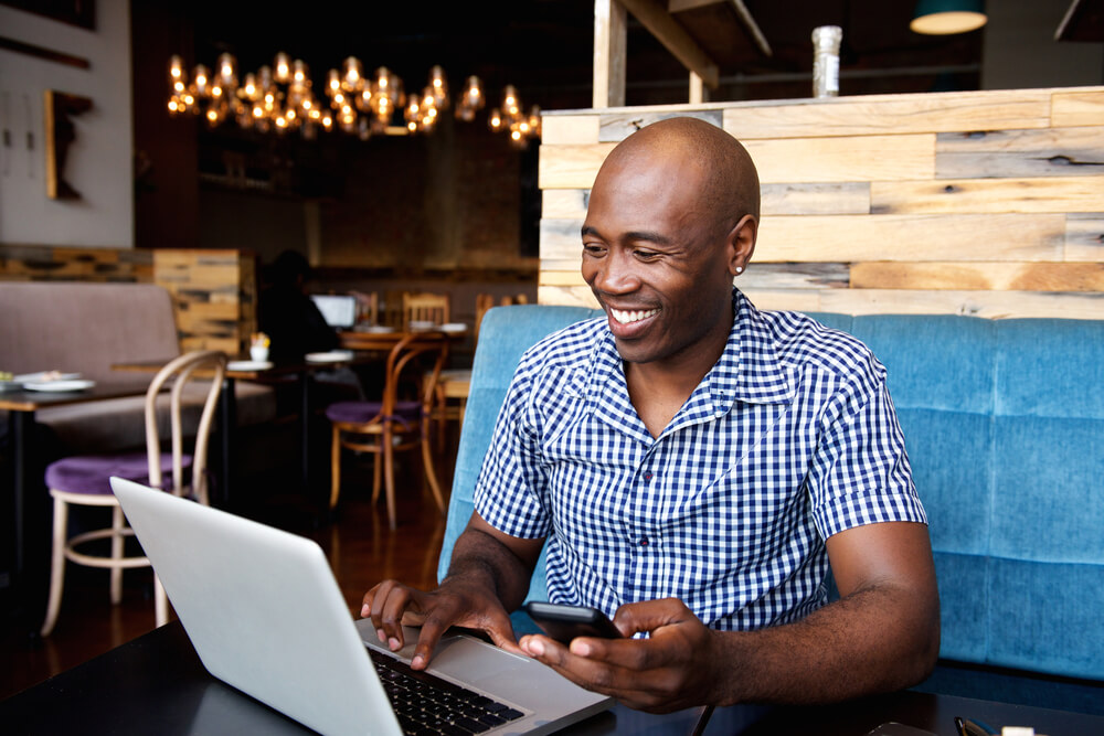 Homem negro e careca sorrindo enquanto usa o notebook e o celular em estabelecimento comercial.