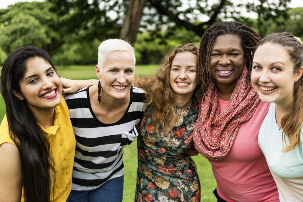 Mulheres negras, brancas, idosas, loiras e morenas reunidas em abraço.